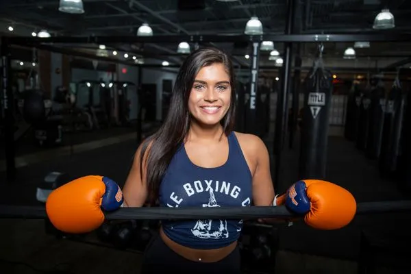 Riley Sanders, Head of Group Classes, smiling athletic woman in a tank top leaning on boxing gloves over a ring rope