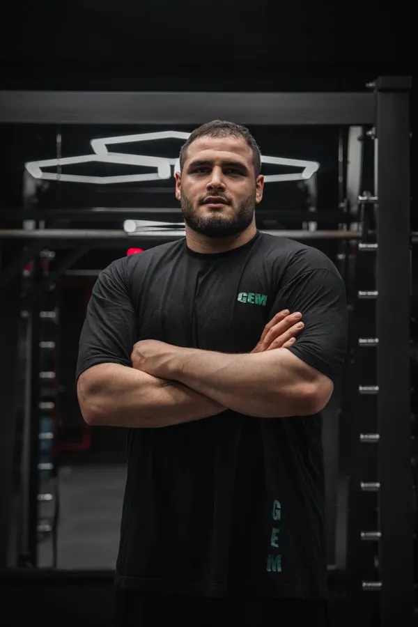 Dre Whitman, Strength Coach, broad bearded muscular man with arms crossed standing in front of a power rack in a dark gym