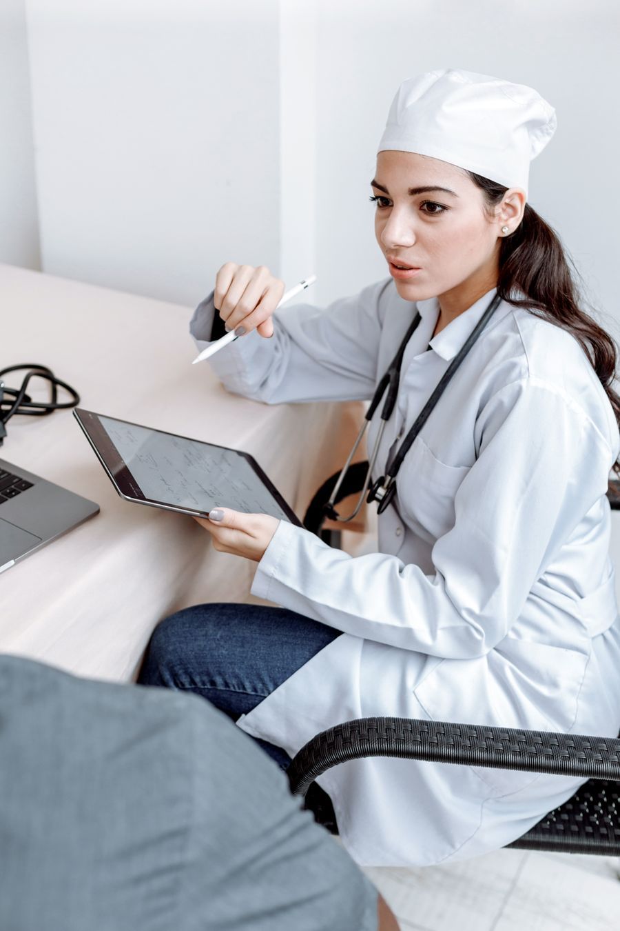 Clinician in a white coat holding a tablet and pointing while speaking with a patient across a desk, representing a one-on-one health coaching consultation.